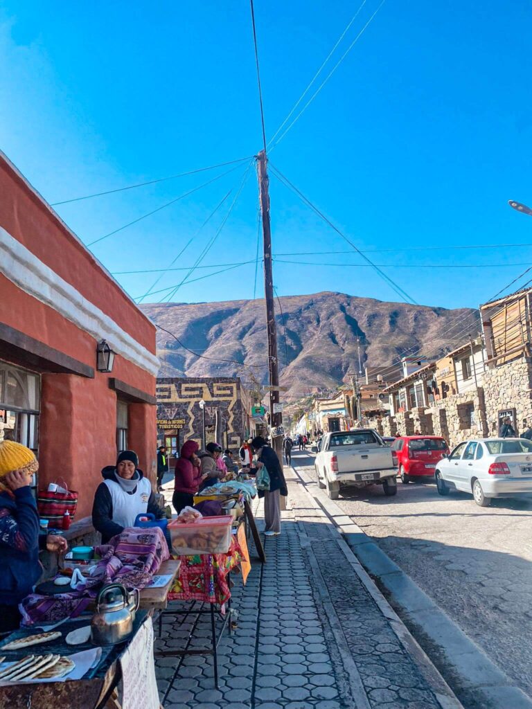 Street scene in Tilcara, a small mountain town, with local women selling food at outdoor tables while cars drive past and mountains rise in the background. Everyday view of local life travelers may experience while visiting smaller towns in Argentina as a solo female traveler.