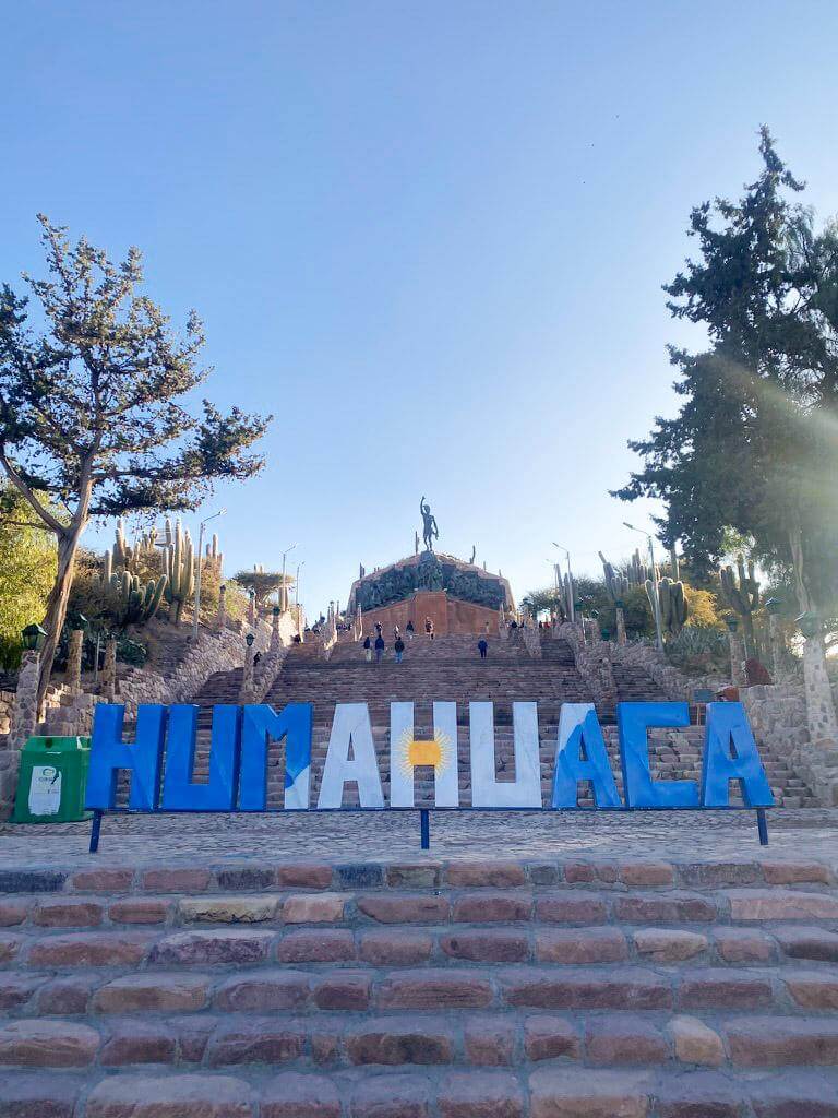 Large blue and white "Humahuaca" sign at the base of stone steps leading up to the Monument to the Heroes of Independence on a hill surrounded by cacti and trees under a clear sky. Iconic landmark visitors see when arriving in the town and exploring whether Humahuaca is worth visiting in northern Argentina.