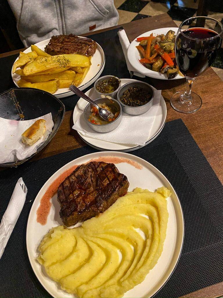 Plates of Argentine steak served with mashed potatoes and thick potato wedges alongside small bowls of chimichurri sauce and a glass of red wine on a restaurant table. Classic Argentine dinner many visitors try during a 2 days in Buenos Aires itinerary.