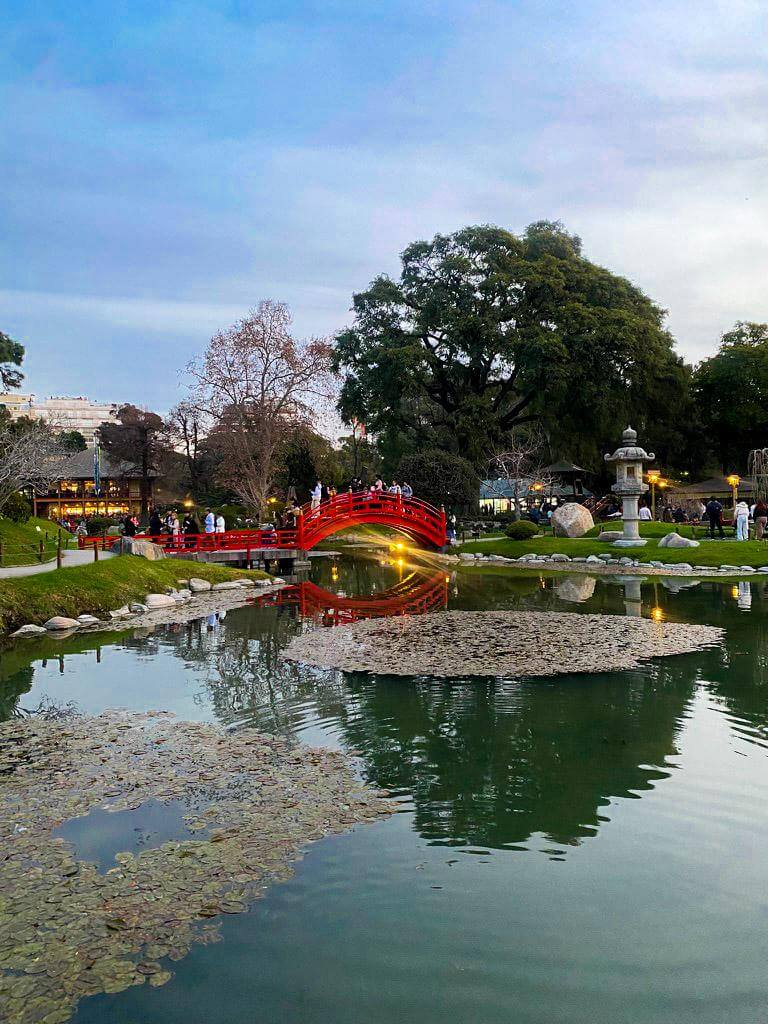 Red arched bridge crossing a pond in the Japanese Garden of Buenos Aires with people walking across it and trees surrounding the water at sunset. Peaceful park setting in Palermo often included as a relaxing stop during a 2 days in Buenos Aires itinerary.