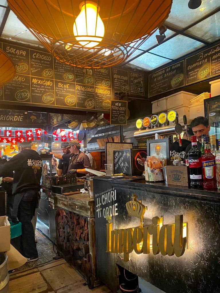 Busy food stall inside a Buenos Aires market where cooks prepare choripán behind the counter beneath a chalkboard menu listing options like "Solapa 6100", "Criollo 5000", "Mexicano 7500", and "Patagónico 8100". The stand labeled "Chori #1" and "Imperial" beer taps shows the lively street food scene many visitors try during a 2 days in Buenos Aires itinerary.