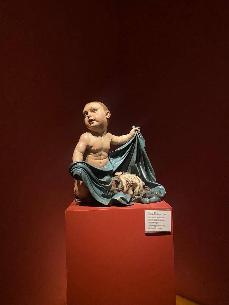 Small sculpture of a child holding a draped cloth displayed on a pedestal inside a museum gallery with deep red walls and dramatic lighting. Historic artwork exhibited in a Buenos Aires museum often included on a cultural stop during a 2 days in Buenos Aires itinerary.