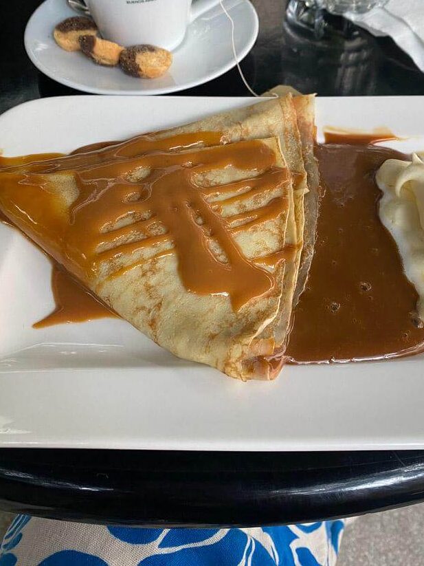 Thin crepe folded and drizzled with dulce de leche served on a white plate with extra sauce and a swirl of cream alongside a cup of tea on a café table.