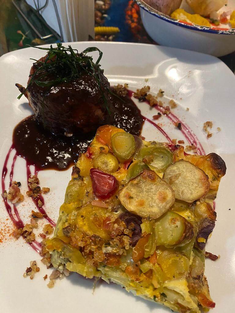 Plated llama meat in a rich dark sauce topped with herbs served alongside a slice of vegetable bake with potatoes and peppers, presented on a white plate in a restaurant setting.