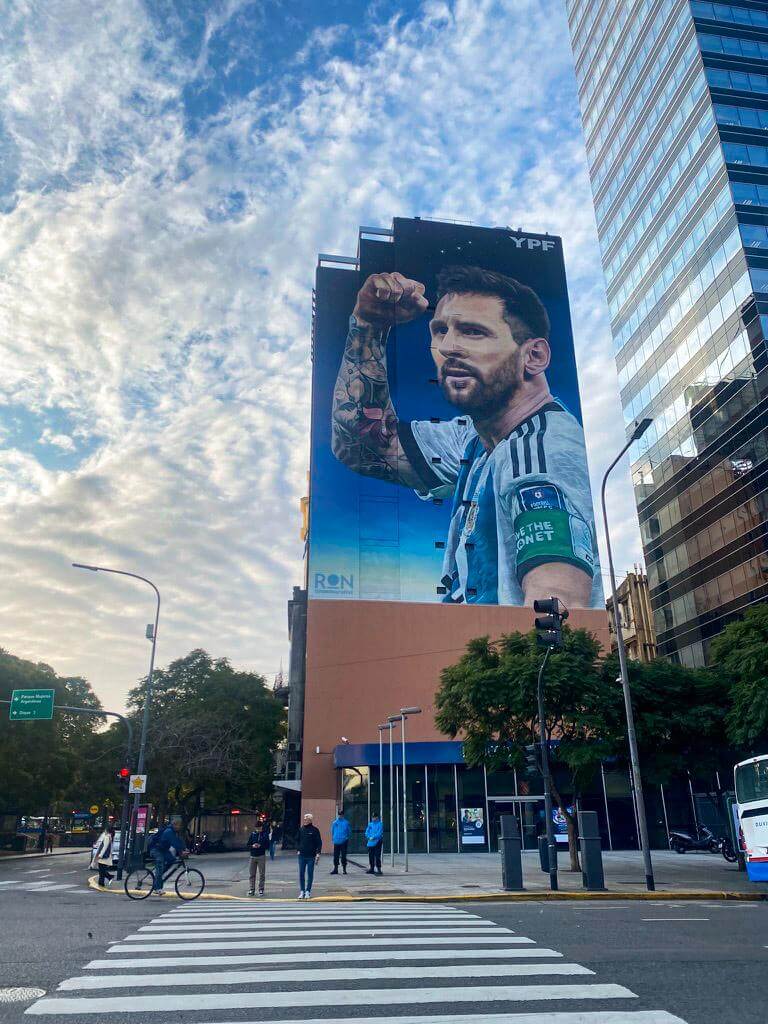Alt text: Large mural of Lionel Messi in an Argentina jersey covering the side of a building in Buenos Aires with people crossing the street below.