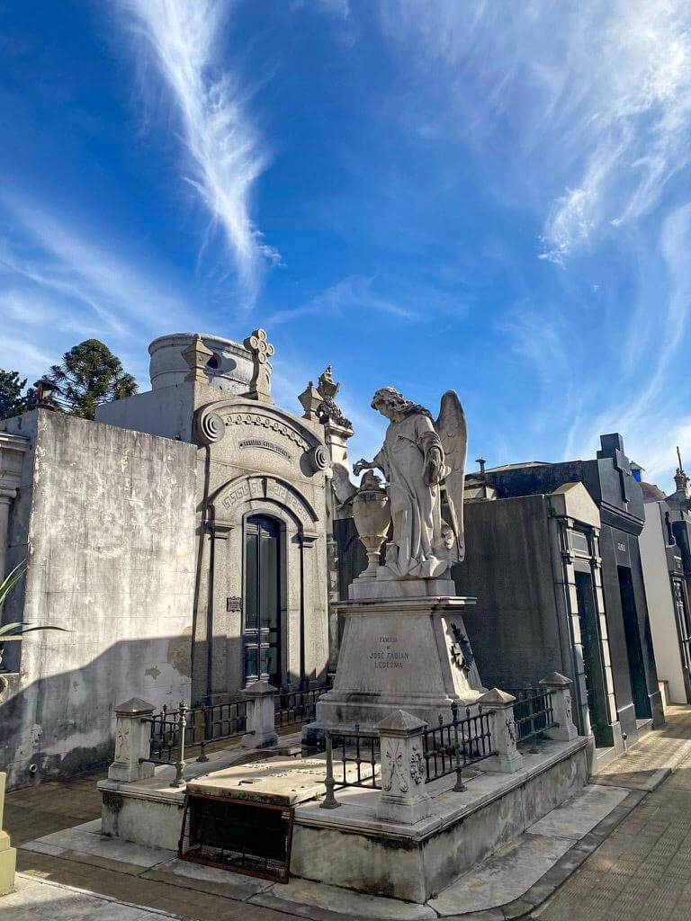 Ornate marble mausoleums and angel statue inside Recoleta Cemetery in Buenos Aires under a bright blue sky with wispy clouds above. Historic cemetery known for elaborate tombs and famous graves often visited during a 2 days in Buenos Aires itinerary.